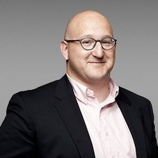 Portrait d'un homme en costume noir et chemise rose, souriant devant un fond neutre.